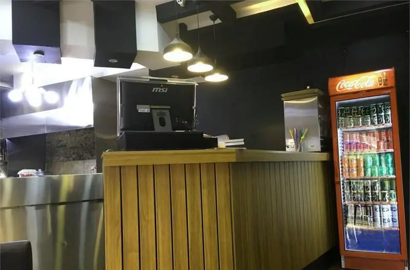 نمای داخلی فست فود ویچنزا با پیشخوان چوبی، مانیتور MSI، یخچال نوشیدنی کوکاکولا پر از قوطی و آشپزخانه استیل مدرن.