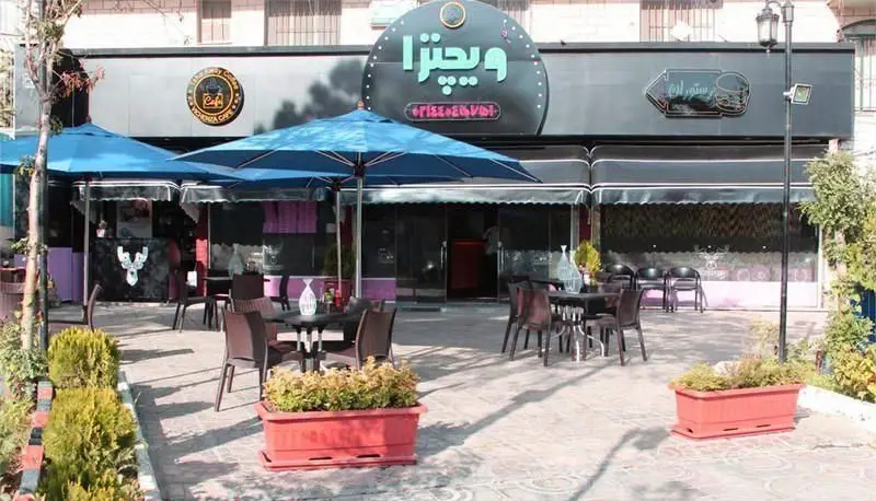 فست فود و کافه ویچنزا با فضای باز و دلنشین، میز و صندلی‌های متعدد، چترهای آبی و گلدان‌های زیبا مناسب پیتزا و غذا.