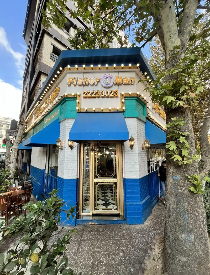 تصویری از نمای بیرونی رستوران دریایی مرد ماهی‌گیر شعبه اندرزگو، ساختمانی گوشه‌ای آبی و سفید با سایه‌بان‌های آبی و فضای نشستن در فضای باز.