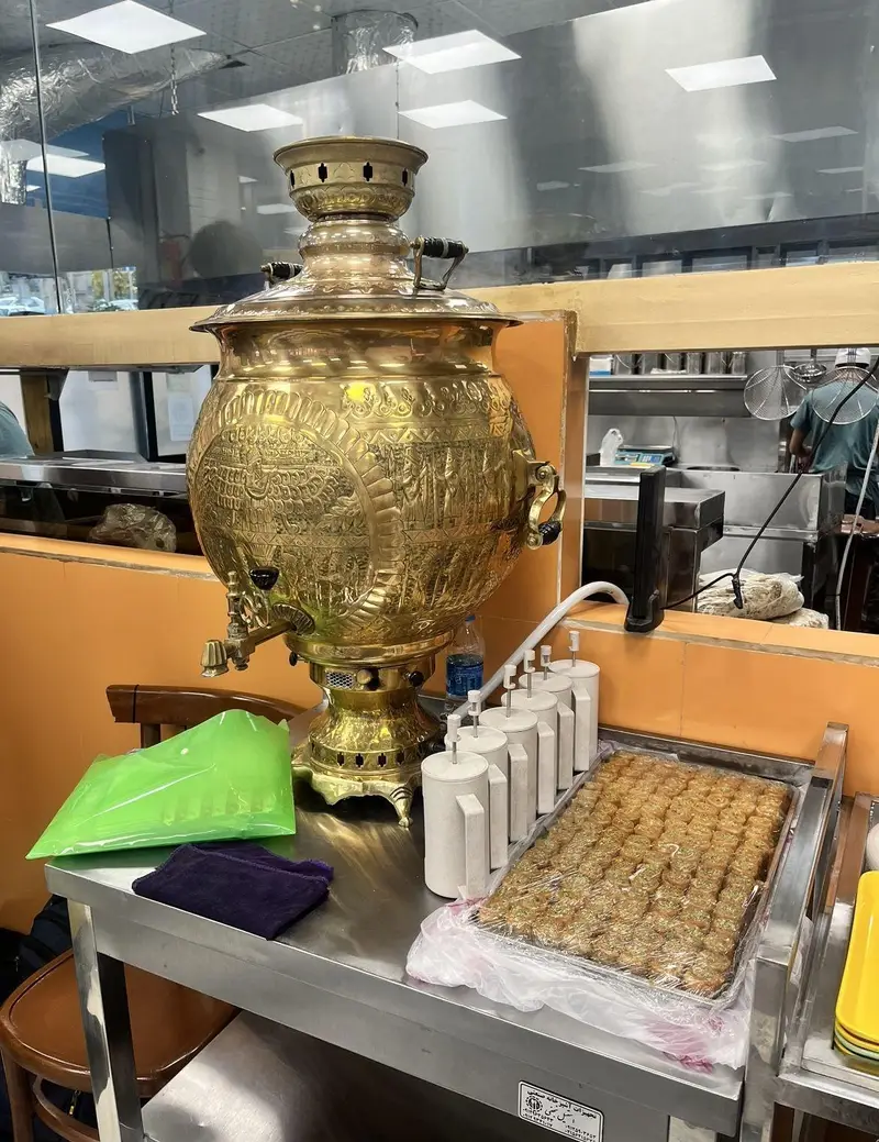 سماور بزرگ برنجی و سینی باقلواهای تازه در رستوران مرد ماهی‌گیر شعبه اندرزگو، فضایی دلنشین برای تجربه چای و دسر سنتی.