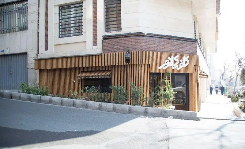 کافه کلاور: تصویری از نمای بیرونی کافه با طراحی چوبی عمودی جذاب، تابلوی نام کافه به رنگ سفید، ورودی شیشه‌ای و فضای سبز مقابل آن در خیابان تهران.