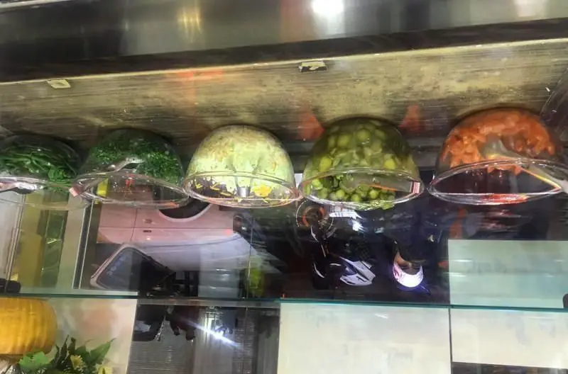 مواد اولیه تازه و متنوع ساندویچی فلافل مامان جون شامل کاهو، سبزی، کلم، خیارشور و سالاد مخصوص در ظرف‌های شیشه‌ای نمایش داده شده است.