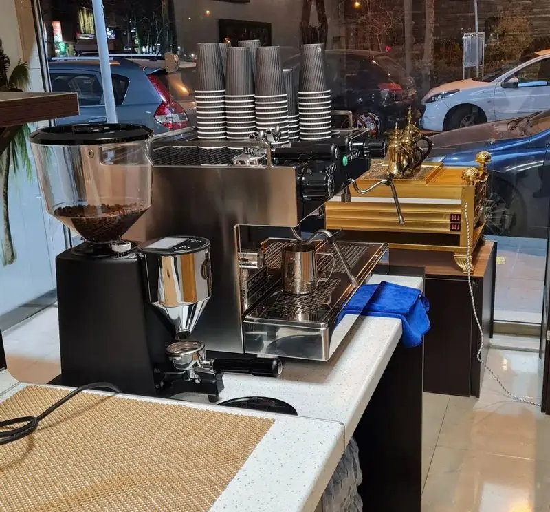 فضای داخلی کافه نخل با دستگاه اسپرسو براق، آسیاب قهوه، فنجان‌های یکبار مصرف و دکوراسیون طلایی. آماده سرو قهوه تازه و خوش عطر.