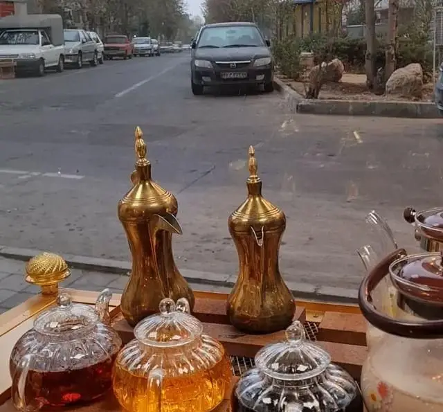 نمایش چشم‌نواز قوری‌های شیشه‌ای چای و ظروف سنتی طلایی در ویترین کافه نخل، منظره‌ای جذاب از شهر