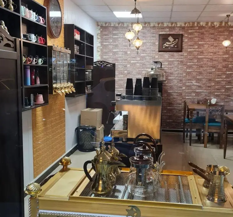 کافه نخل: فضایی دلنشین با دکور آجری، دستگاه قهوه ساز، قفسه‌های فنجان و دله‌های سنتی، میز و صندلی‌های چوبی برای مشتریان.