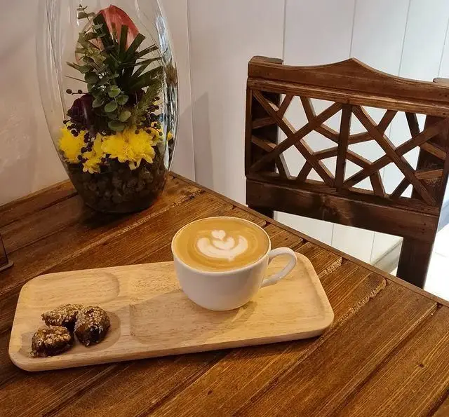 تصویری دلنشین از یک فنجان لاته آرت با طرح قلب و سه شیرینی کنجدی بر روی سینی چوبی در کافه نخل، فضایی گرم و دوستانه.