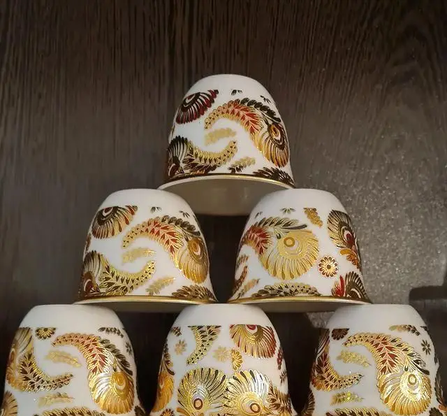 شش فنجان قهوه و چای نفیس سفید با نقوش پیچیده طلایی، قرمز و مشکی، چیده شده به صورت هرمی، ایده‌آل برای سرو در کافه نخل.