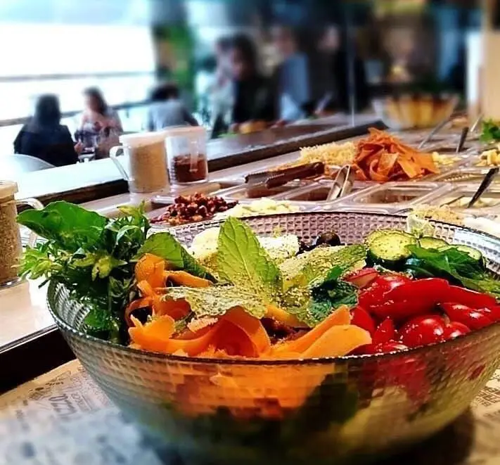 کاسه سالاد تازه و رنگارنگ در پیش‌زمینه و سالاد بار بونو ظفر پر از سبزیجات متنوع و مواد اولیه باکیفیت برای غذای سالم در رستوران ایتالیایی.