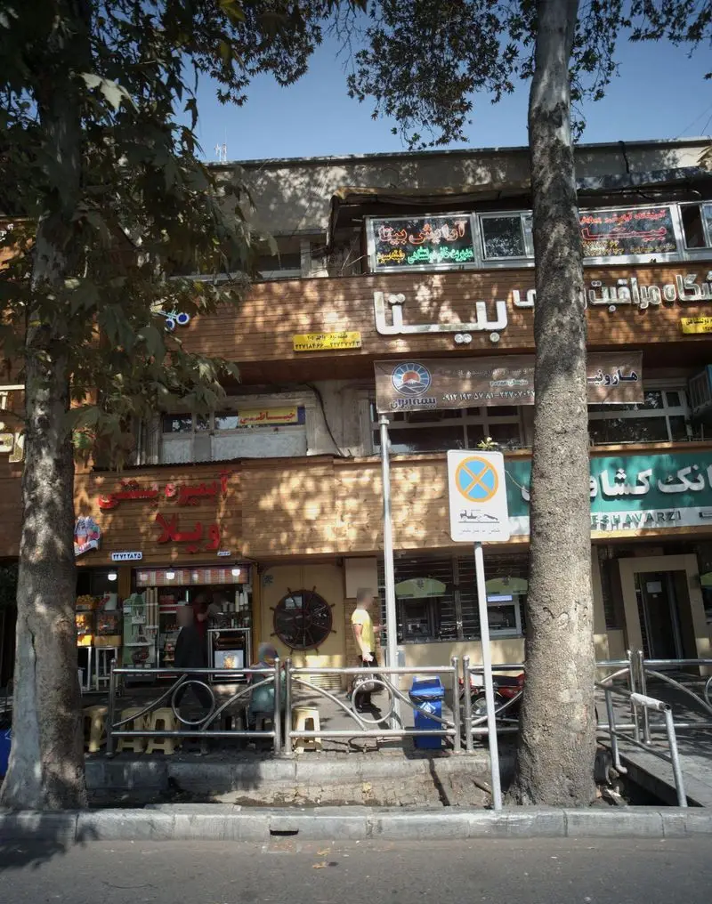 تصویر آبمیوه بستنی ارگ در یک خیابان شلوغ شهری با درختان بلند، مردم در حال خرید و تابلوهای رنگی مغازه های اطراف و بانک کشاورزی.