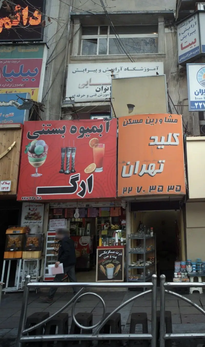 آبمیوه بستنی ارگ با تابلوهای رنگی انواع بستنی، آبمیوه تازه و ذرت مکزیکی در نمای یک خیابان پرتردد شهری.