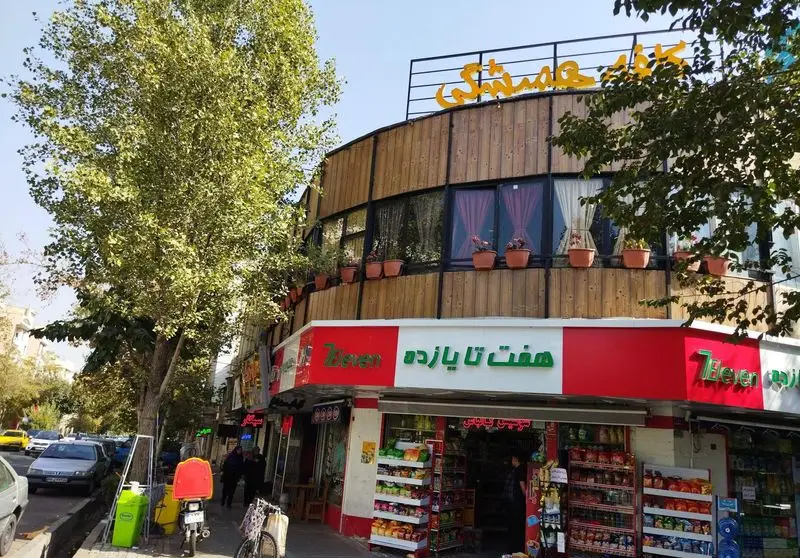 نمای بیرونی کافه همیشگی با تابلوی زرد رنگ و دکور چوبی، گلدان‌ها و پنجره‌های بزرگ، بالای سوپرمارکت هفت تا یازده در خیابان.