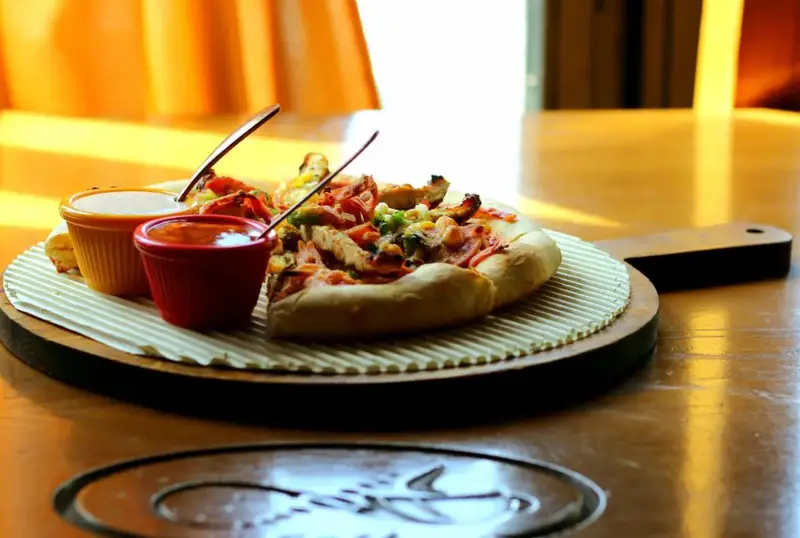 پیتزای تازه و خوش‌طعم کافه همیشگی با سس‌های ویژه سرو شده روی تخته چوبی، تجربه‌ای دلپذیر از یک وعده غذایی کامل.