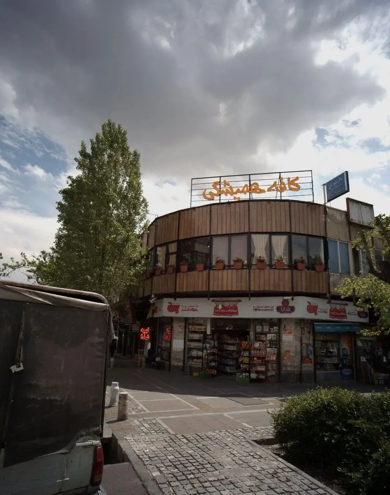 تصویری از کافه همیشگی با تابلو زرد رنگ بر فراز ساختمانی دایره‌ای شکل، نمای چوبی، پنجره‌ها، فروشگاه در همکف و آسمان ابری.