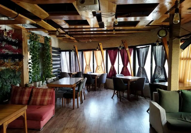 فضای داخلی دنج و زیبای کافه همیشگی با سقف چوبی، مبلمان متنوع، دیوار سبز و پنجره‌های رنگی، ایده‌آل برای دورهمی و استراحت.