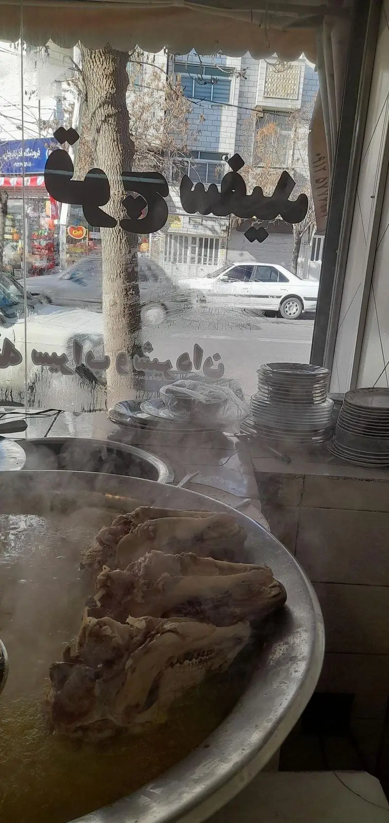 تصویری از نمای داخلی طباخی قوچ سفید، دیگ‌های بزرگ کله‌پاچه در حال طبخ با بخار آب گرم، ظروف آماده سرو، و منظره خیابان از پنجره.