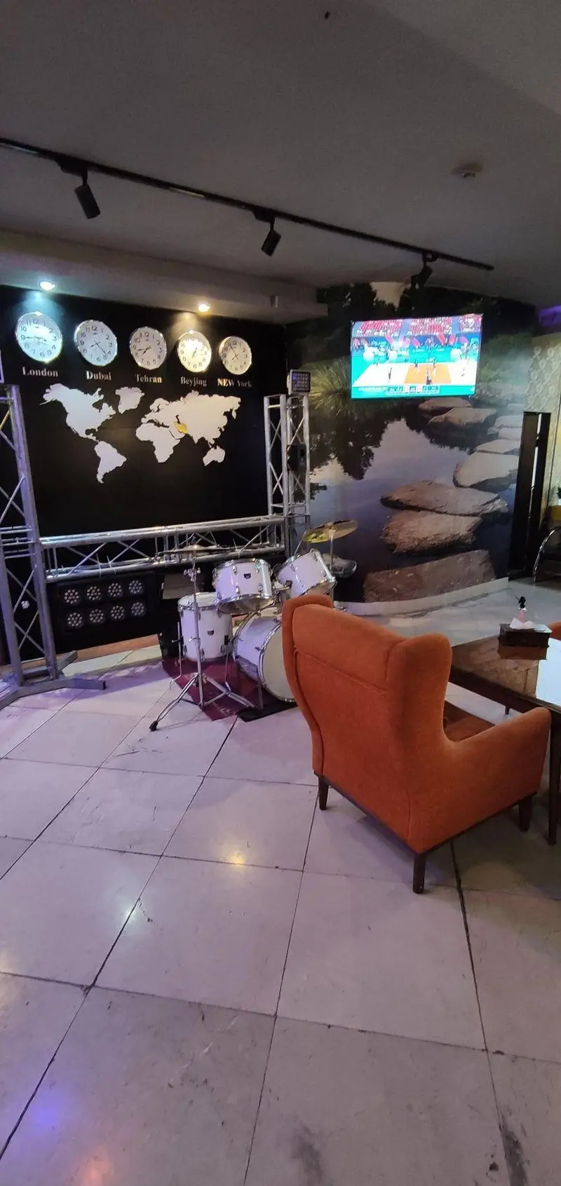 محیط داخلی کافه رستوران سالار با طراحی مدرن، نقشه جهان، ساعت شهرهای مهم، درام ست، تلویزیون بزرگ و صندلی‌های دنج نارنجی.