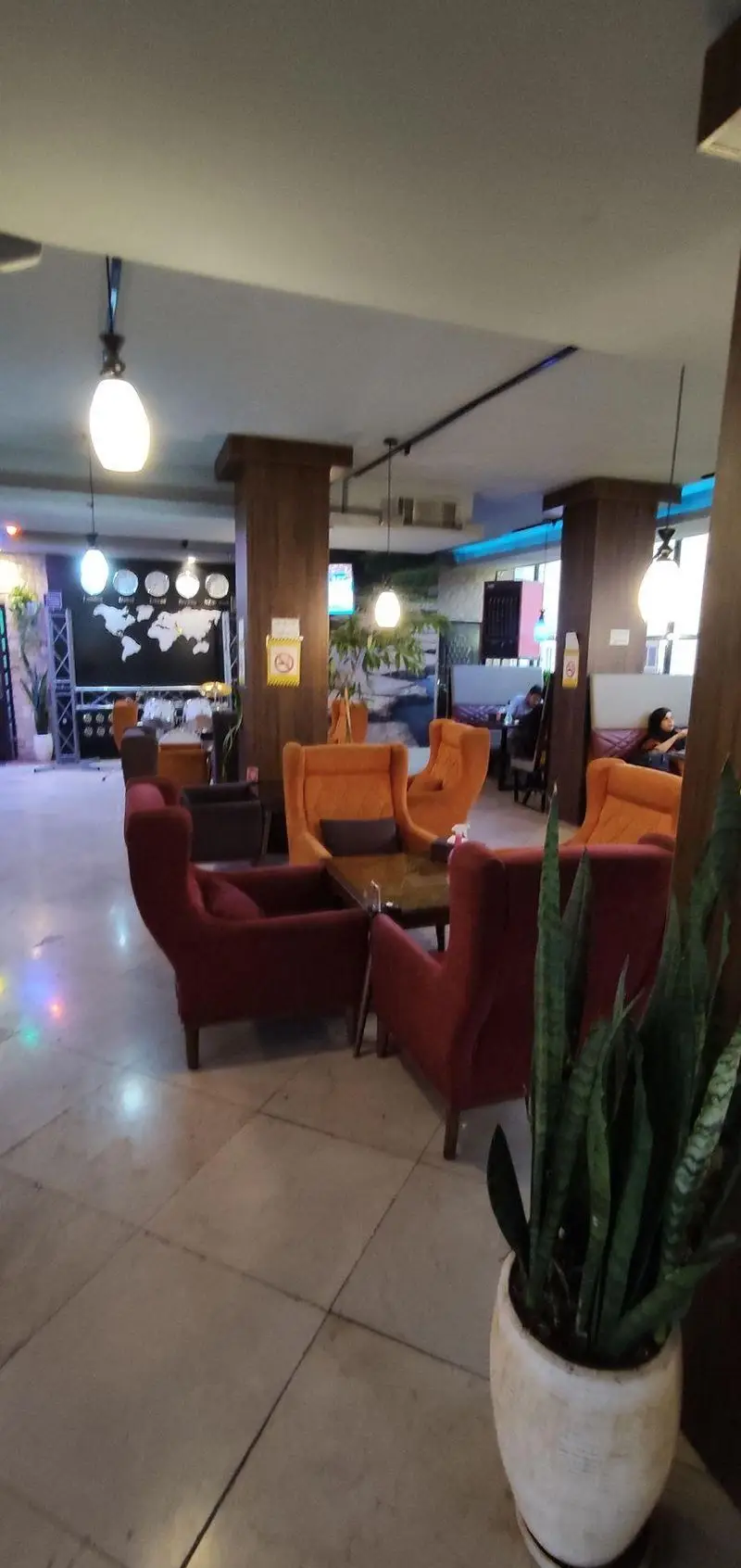 کافه رستوران سالار: فضای داخلی آرام و شیک با صندلی‌های راحتی قرمز و نارنجی، میزها، دکوراسیون مدرن، نقشه جهان و گیاهان تزئینی.