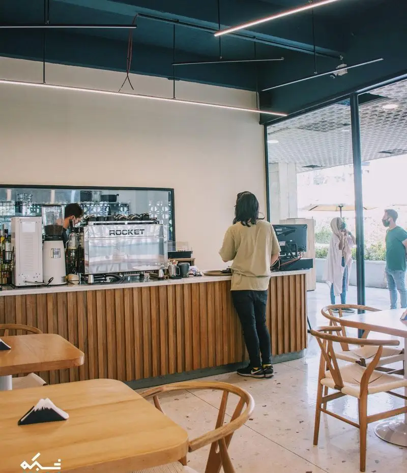 کافه یارد، فضای داخلی مدرن و دلنشین، باریستا پشت کانتر چوبی با دستگاه اسپرسو راکت، دکوراسیون شیک و میز و صندلی‌های چوبی. محیطی جذاب برای قهوه‌دوستان.