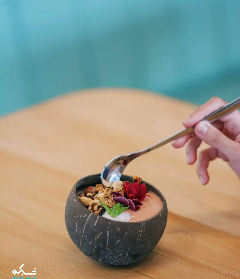 یک کاسه اسموتی نارگیلی زیبا و سالم از کافه یارد، مزین به گرانولا و گل، که با قاشق آماده سرو است. تجربه‌ای دلپذیر و مقوی.