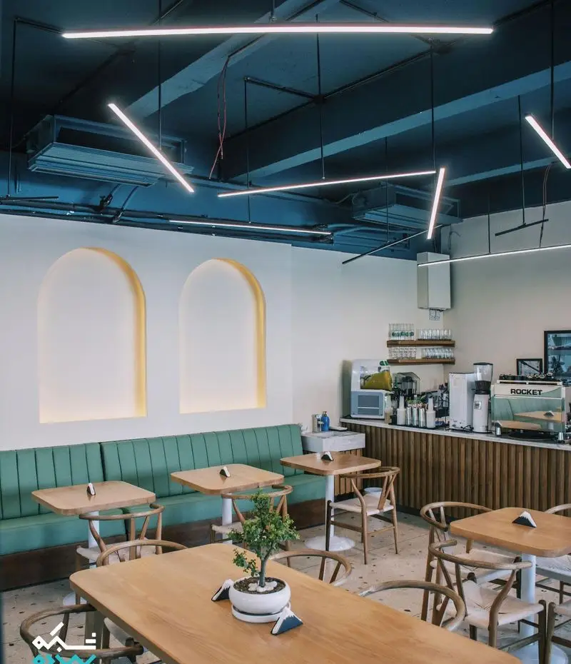 فضای داخلی کافه یارد با دکوراسیون مدرن و شیک شامل میز و صندلی چوبی، مبلمان راحتی سبز، سقف تیره با نورپردازی خطی و دیوار سفید با طاقچه‌های نورانی.