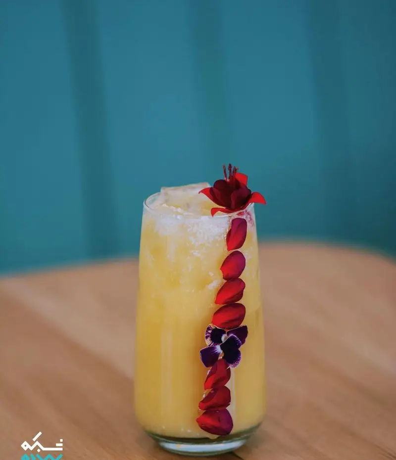 نوشیدنی زرد و دلپذیر کافه یارد، تزیین شده با گلبرگ‌های قرمز و گل بنفش روی میز چوبی کافه. تجربه‌ای خنک و تازه.