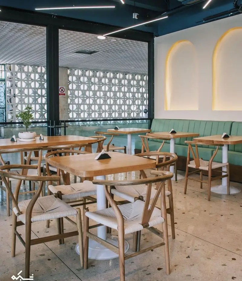 دکوراسیون داخلی کافه یارد با میز و صندلی چوبی مدرن، مبل راحتی سبز، و پنجره‌های بزرگ با نمای بلوک‌های تزئینی.