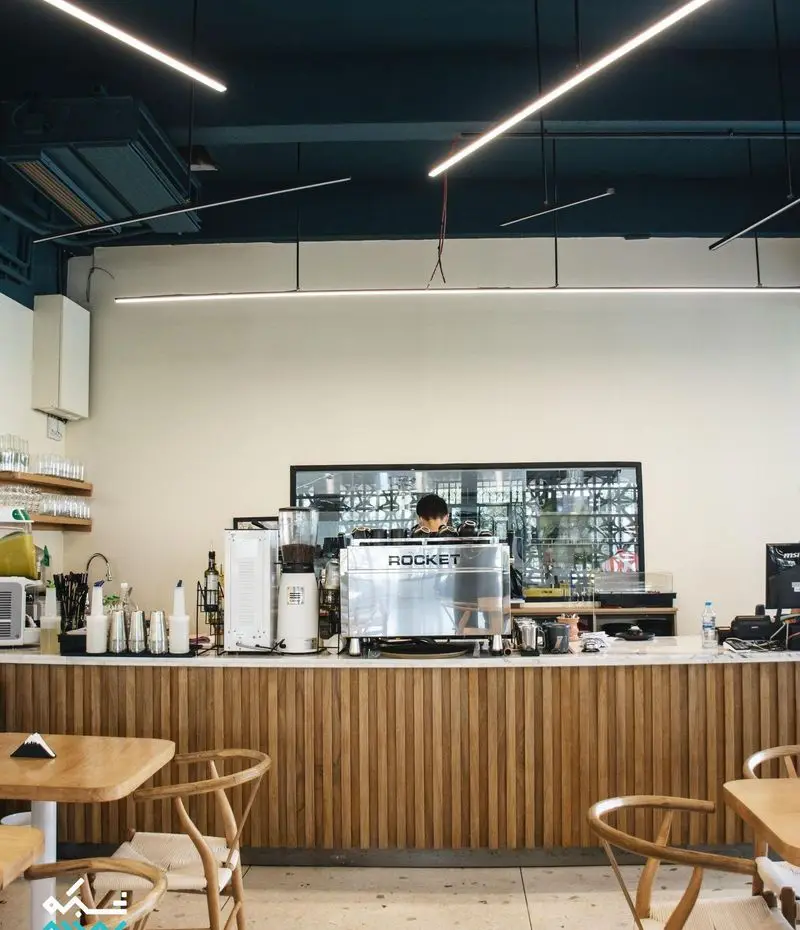 فضای داخلی مدرن کافه یارد با دکوراسیون چوبی، پیشخوان اصلی مجهز به دستگاه اسپرسو راکت و نورپردازی خاص، محیطی دنج و آرام.