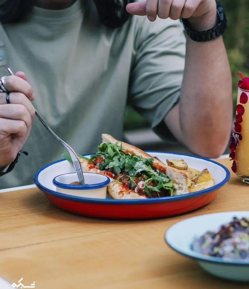 ساندویچ گوشت و آروگولا با سس و سیب‌زمینی در بشقاب قرمز و سفید روی میز چوبی کافه یارد که توسط مشتری در حال میل شدن است.