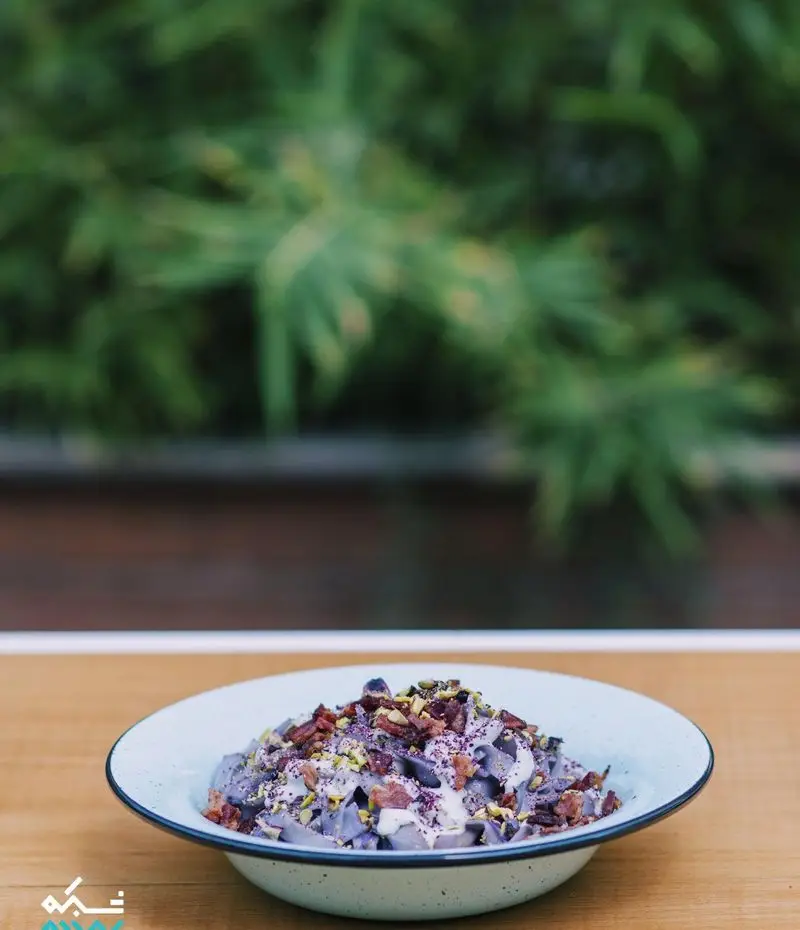 پاستای بنفش خاص کافه یارد با پسته و دانه‌های قرمز تیره، همراه با سس سفید، روی میز چوبی. تصویری دلنشین از منوی خلاقانه کافه یارد.