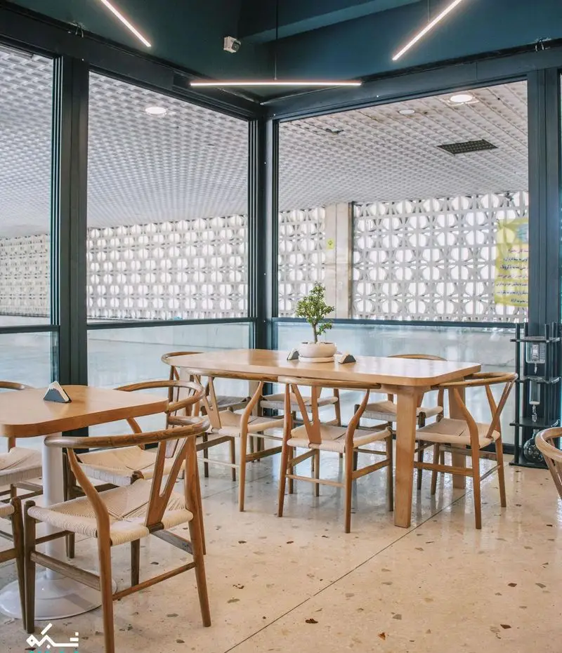 طراحی داخلی زیبا و دنج کافه یارد با میزهای چوبی، صندلی‌های حصیری، کفپوش روشن و چشم‌انداز بیرون از پنجره‌های بزرگ.