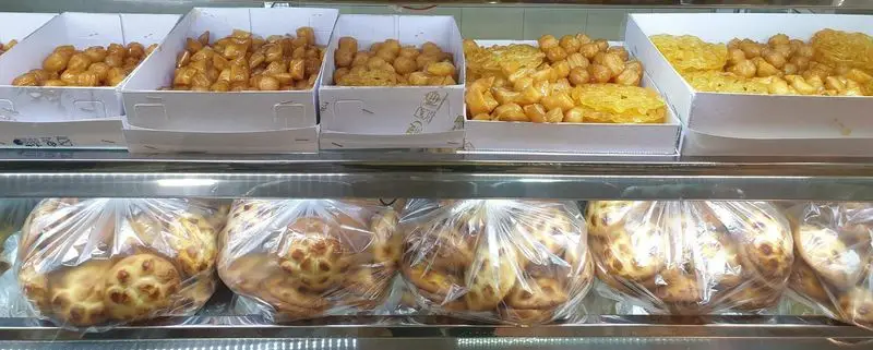 تصویری نزدیک از ویترین پربار قنادی فرح بخش با ردیفی از شیرینی‌های طلایی رنگ سنتی مانند زولبیا، بامیه و همچنین نان‌های محلی تازه و بسته‌بندی شده در پایین، آماده عرضه به مشتریان.