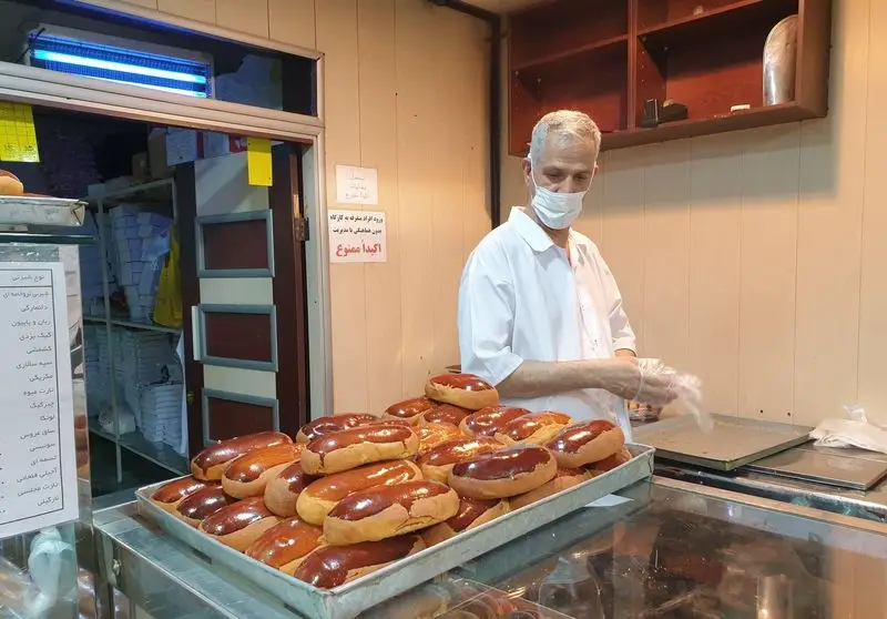 قنادی فرح بخش: مرد قناد با ماسک و دستکش در حال آماده سازی نان شیرینی‌های تازه پخت و براق در محیطی بهداشتی و تمیز.