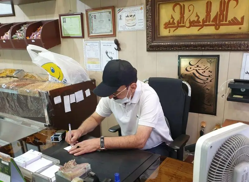 تصویری از صاحب یا کارمند قنادی فرح بخش در حال مدیریت امور، در دفتر مغازه با ویترین‌های پر از خشکبار و شیرینی‌جات.