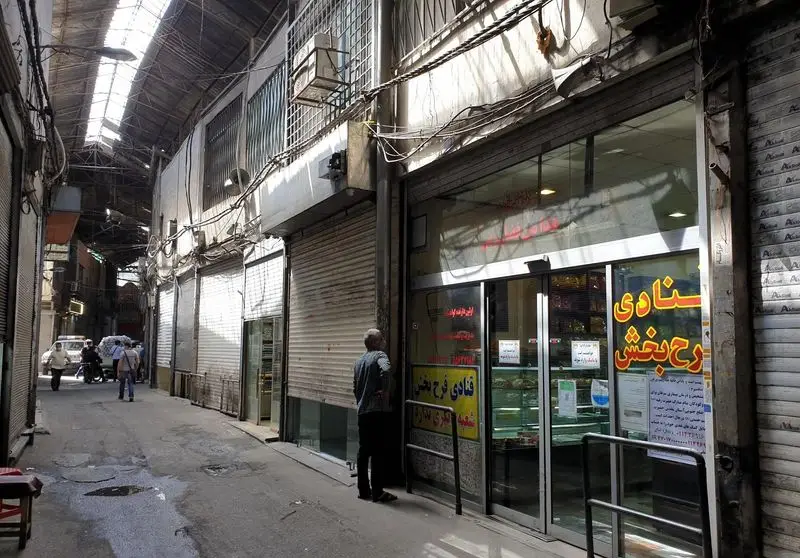 تصویر کوچه ای در بازار ایران با مغازه قنادی فرح بخش و ویترین شیشه‌ای پر از انواع شیرینی‌های سنتی و مدرن در یک روز آفتابی.