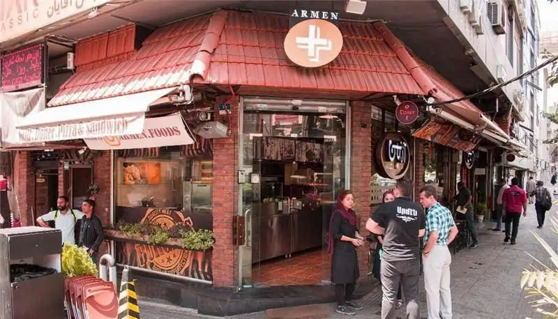 نمای بیرونی پر جنب و جوش فست فود مجموعه آرمن با تابلو Doner, Pizza & Sandwich و لوگوی متمایز پلاس. مردم در حال گپ و گفتگو.