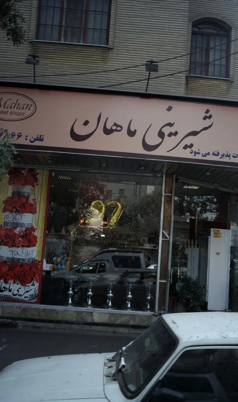 نمای کامل از شیرینی ماهان، یک قنادی و فروشگاه کیک، با تابلوی خوانا، ویترین شیشه‌ای بزرگ، دکور کیک چند طبقه و بالون عدد ۲۷