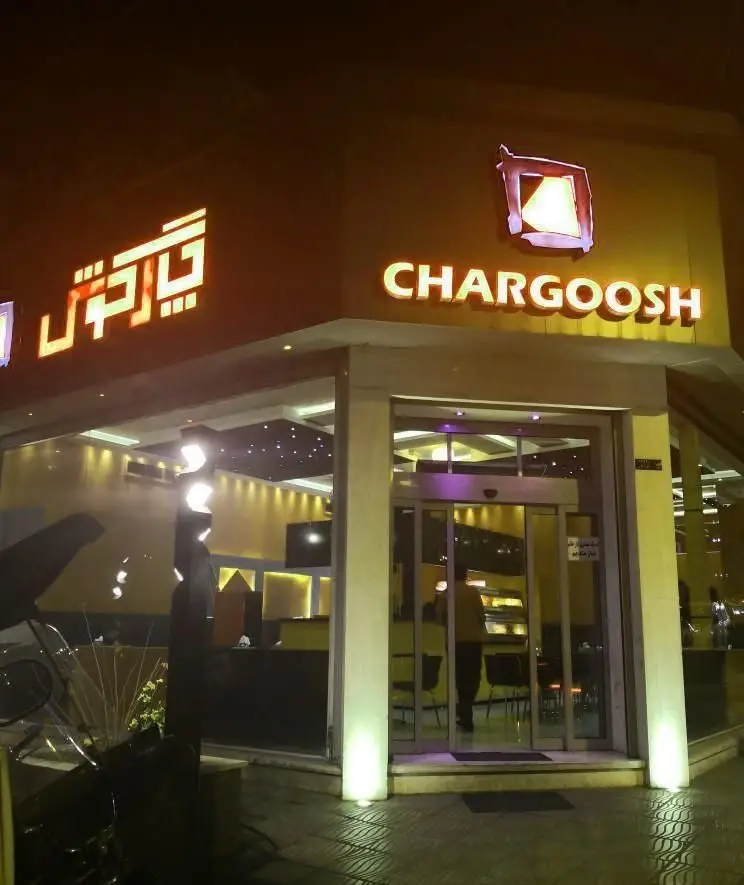عکس شب از فست فود چارگوش با تابلوهای نئون درخشان CHARGOOSH و نام فارسی آن. ورودی شیشه‌ای رستوران نورپردازی شده است.