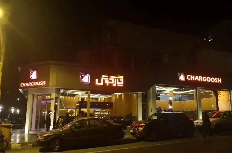 نمای بیرونی فست فود چارگوش در شب، با تابلوهای نورانی CHARGOOSH و متن فارسی "چارگوش"، همراه با فضای داخلی روشن و خودروهای پارک شده در خیابان.
