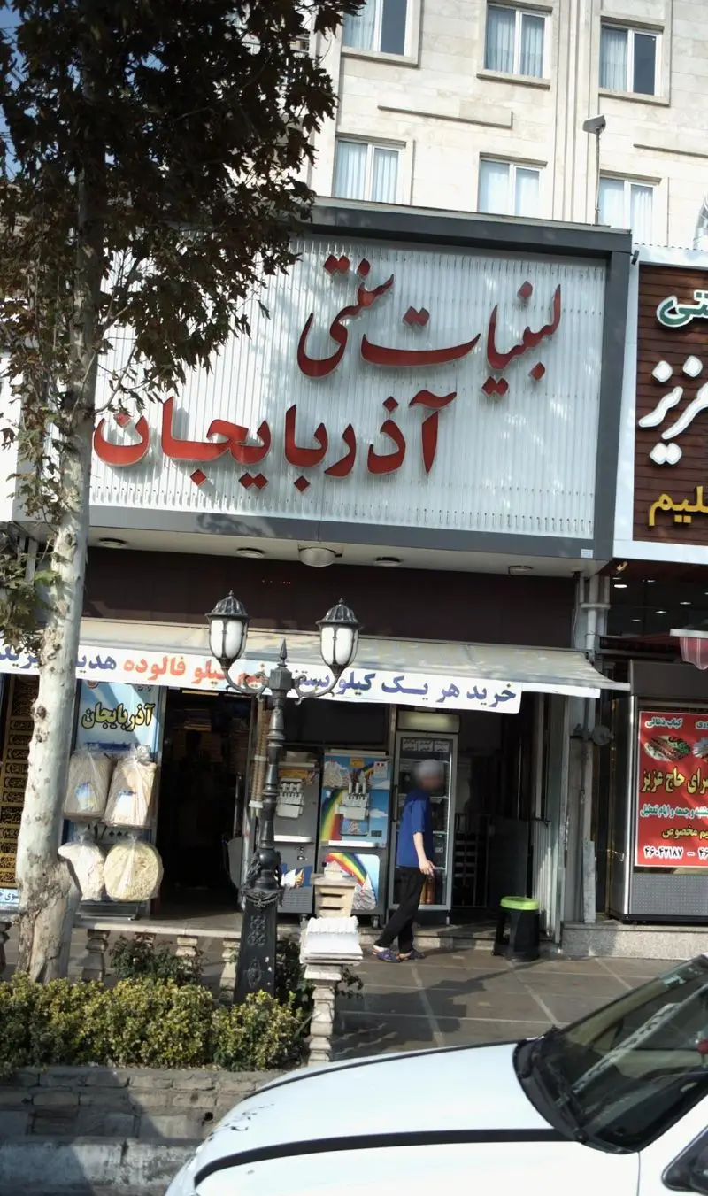 فروشگاه لبنیات سنتی آذربایجان: تابلوی برجسته قرمز، محصولات لبنی و نان تازه، نمای شهری. مناسب برای خرید سنتی.