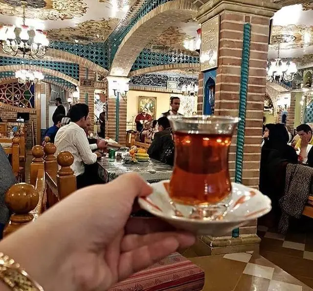 فضای داخلی سفره خانه سنتی باربد با سقف‌های گچ‌بری، آجرکاری‌های فیروزه‌ای و سرو چای در محیطی گرم و اصیل ایرانی.