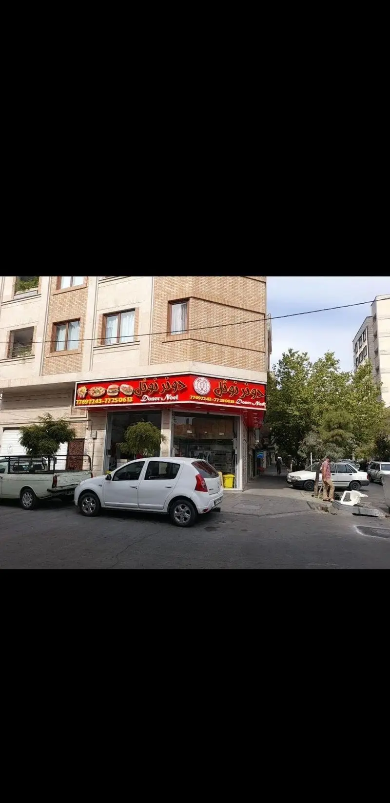 فست فود دونر نوئل، نمایی از مغازه در تقاطع خیابان با تابلوی قرمز رنگ و تصاویر غذا، شامل پیتزا، برگر و دونر، محیط شهری.