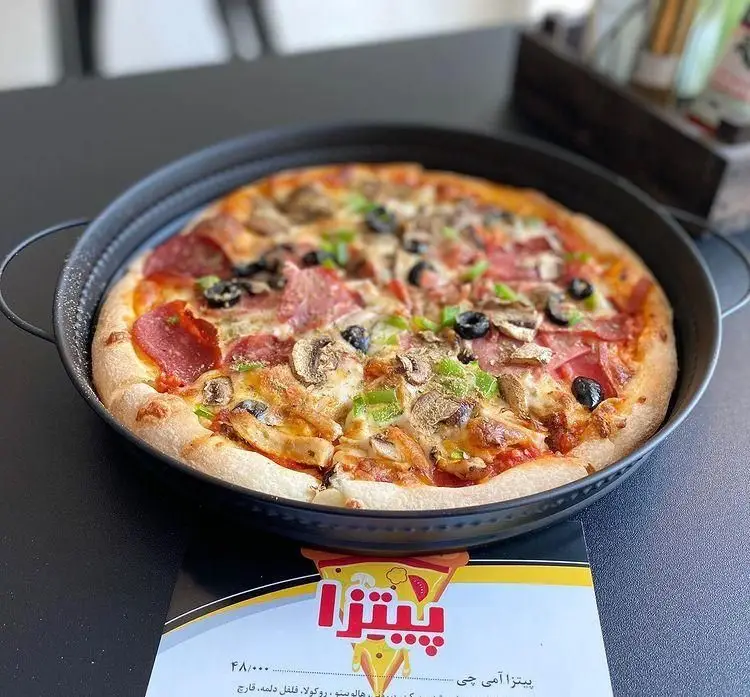 تصویری از پیتزا بزرگ و لذیذ "پیتزا آمی چی" با خمیر تازه، پر از پپرونی، ژامبون، قارچ، زیتون و فلفل دلمه سبز در سینی مخصوص سرو شده.