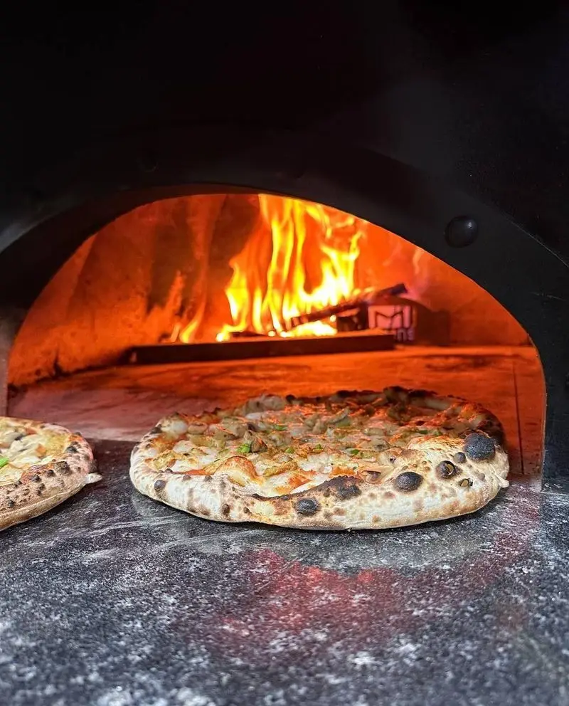 پیتزا داغ و خوشمزه آمی چی در حال پخت داخل فر هیزمی با آتش سوزان، آماده سرو برای مشتریان عزیز در محیطی دلنشین.