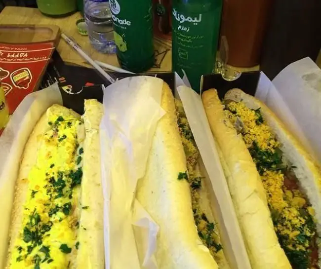 چند ساندویچ هات داگ مسعود تازه و خوشمزه با مخلفات رنگارنگ، در کنار نوشابه لیموناد زمزم خنک. غذایی دلپذیر برای مشتریان.