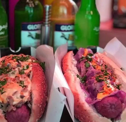 تصویری جذاب از دو هات داگ خاص و خوشمزه از هات داگ مسعود، شامل سوسیس بنفش و مخلفات متنوع روی نان تازه، همراه با نوشیدنی‌ها.