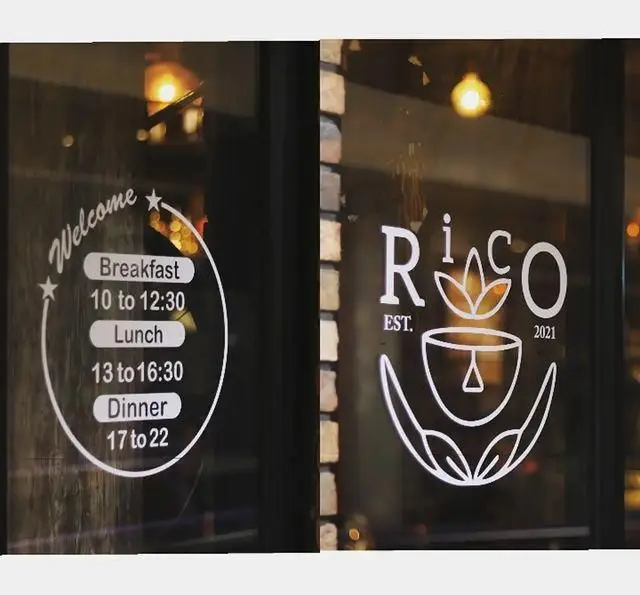پنجره کافه ریکو با دکوراسیون شیک، شامل لوگو Rico EST. 2021 و ساعات کاری صبحانه (10-12:30)، ناهار (13-16:30) و شام (17-22).