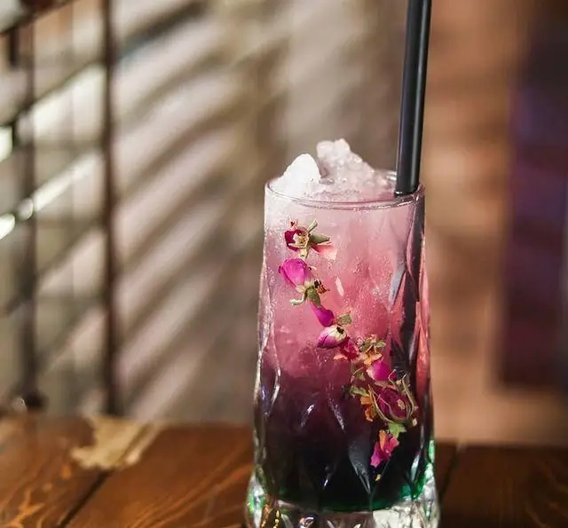 نوشیدنی خنک و چشم‌نواز کافه ریکو: کوکتل لایه‌ای صورتی و بنفش با یخ خرد شده، مزین به غنچه‌های رز خشک و نی مشکی.