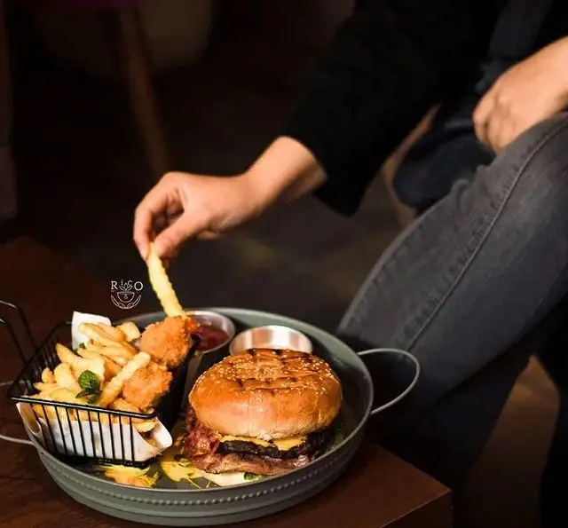 مشتری در کافه ریکو در حال میل کردن همبرگر ویژه با سیب‌زمینی سرخ‌شده و سس در محیطی دوستانه و دلپذیر.