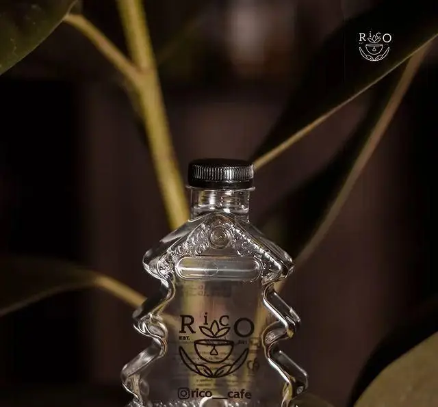 بطری شیشه‌ای منحصر به فرد به شکل درخت از کافه ریکو، حاوی نوشیدنی شفاف. لوگوی "Rico Cafe EST. 2021" و طرح فنجان قهوه روی بطری.