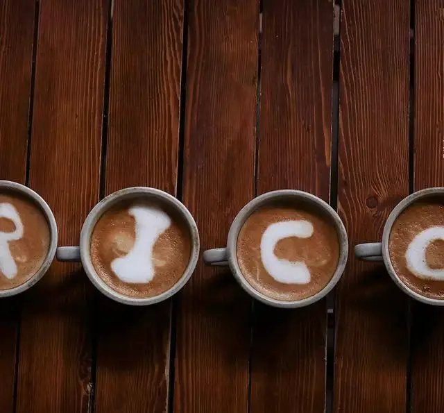 تصویر چهار فنجان قهوه با لاته آرت زیبای RICO روی میز چوبی کافه ریکو، نشانگر کیفیت بالای نوشیدنی‌ها و هنر باریستا.